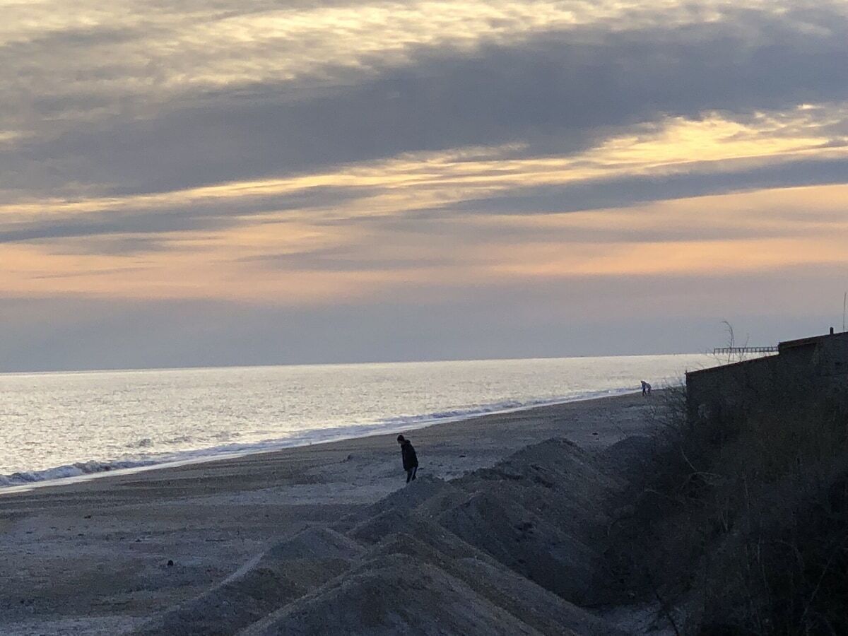 A beach with a person walking along the shore, a sandy area, and a structure on the right.