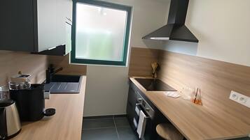 Een kleine keuken met een raam, een kookplaat, een spoelbak en een barstoel.