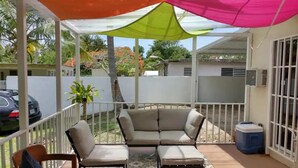 A covered outdoor seating area with a sofa and chairs, a patterned rug, and a blue trash bin.