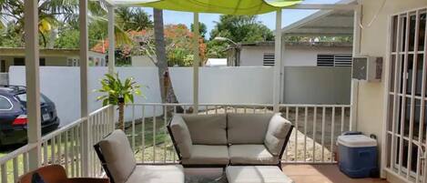 A covered outdoor seating area with a sofa and chairs, a patterned rug, and a blue trash bin.
