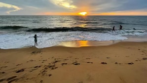 Une plage où l’on voit des empreintes de pas menant à l’eau, un coucher de soleil en arrière-plan, et des gens qui profitent de la plage.