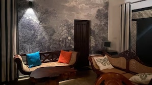 Une pièce avec un plafond haut, un mur à texture, un banc en bois, deux canapés avec des coussins, une porte et une fenêtre avec des rideaux.
