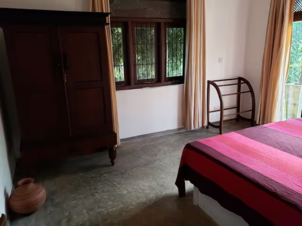 A bedroom with a bed, a wooden wardrobe, a window with curtains, and a small table.