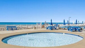 Zona de piscina con sillas reclinables azules y sombrillas, una valla blanca y vista despejada al océano.