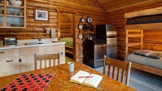 Ein gemütliches Blockhaushaus-Interieur mit einem Holzesstisch, Stühlen, einer Küchenzeile mit Kühlschrank und einem Schlafschlater.