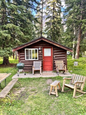 A rustic cabin with a red door, two wooden chairs, a small table, and a grill outside.