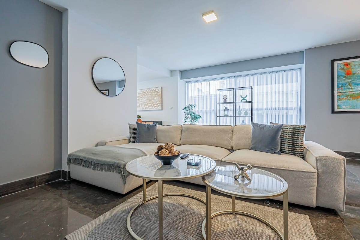 A modern living room with a beige sectional sofa, two glass coffee tables, a round rug, a round mirror, and a window with blinds.