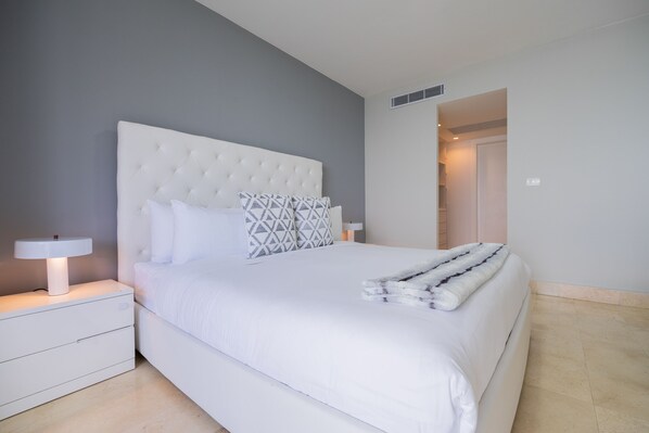 Una cama bien tendida con ropa de cama blanca y un edredón estampado, una mesita de noche con una lámpara y una pared gris.