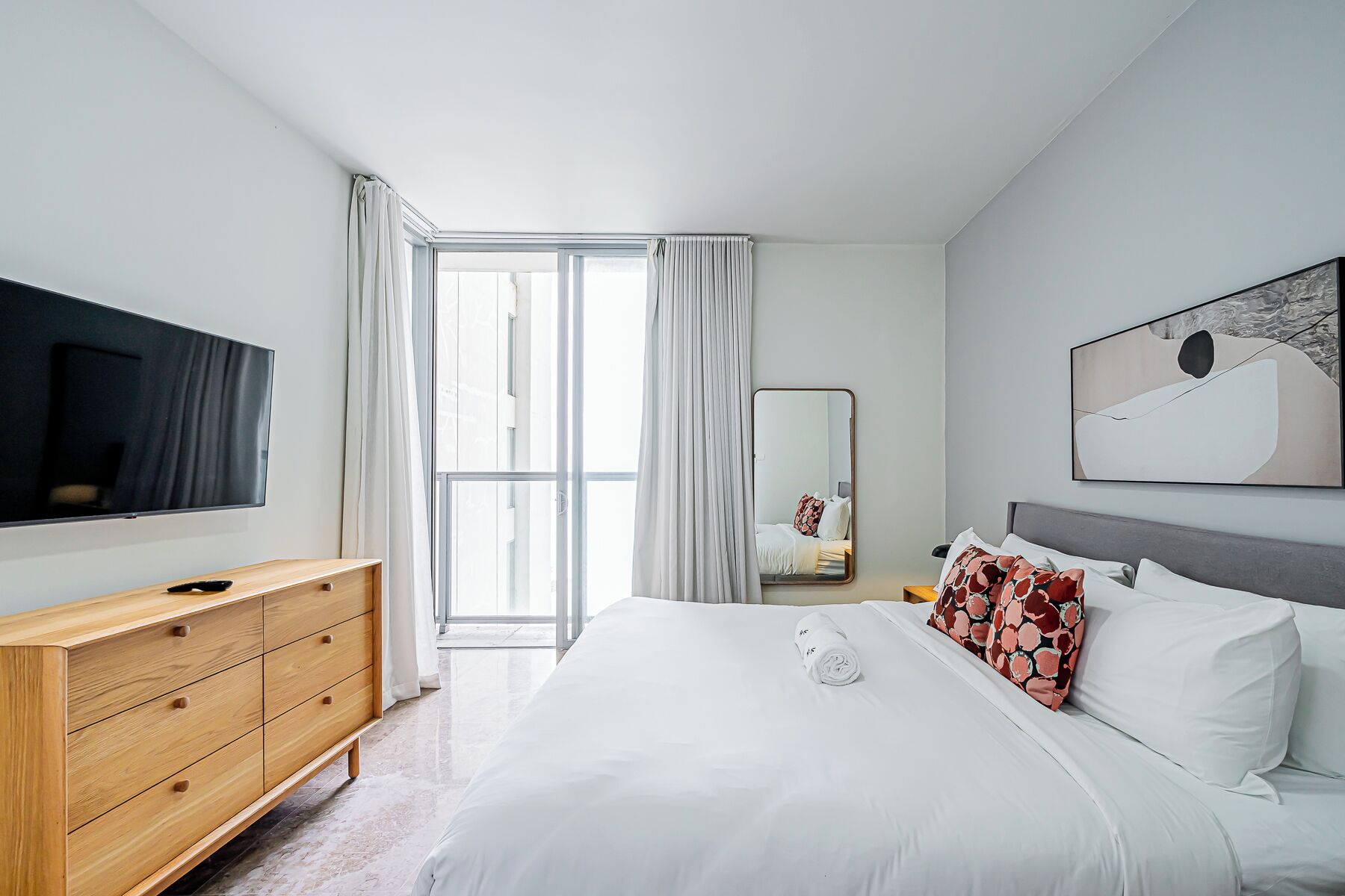 A modern hotel room with a large bed, a wooden dresser, a flat-screen TV, and a large window with curtains.