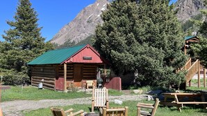 A rustic log cabin with a green roof, surrounded by a grassy area with wooden benches. - Comfy cabin is situated on the Silver Gate property which overlooks 10,000ft tall mountain peaks surrounding the town. (SILVER GATE)