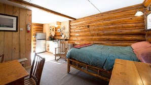 A log cabin room with a bed, a wooden table, a chair, a kitchenette, and a framed picture on the wall. - Comfy cabin is situated on the Silver Gate property which overlooks 10,000ft tall mountain peaks surrounding the town. (SILVER GATE)