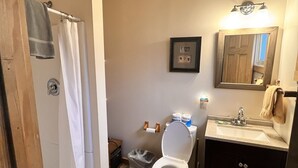 A bathroom with a wooden ceiling, a white toilet, a sink with a mirror, and a shower curtain. - Newly Remodeled Motel Room with 1 Queen Bed, Bathroom with shower. Mini Fridge, Coffee Maker. Situated close to the General Store in Silver Gate, MT (SILVER GATE)