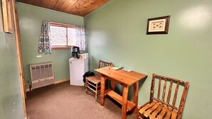 A compact room with a wooden table, two chairs, a small kitchenette, and a window with curtains.