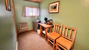 Une petite pièce chaleureuse et accueillante, avec une table en bois, deux chaises, une fenêtre avec des rideaux, un radiateur et un tableau encadré accroché au mur.
