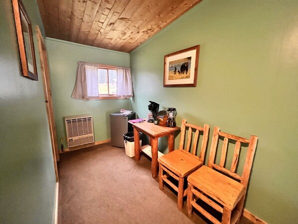 Een knus, klein kamertje met een houten tafel, twee stoelen, een raam met gordijnen, een radiator en een lijst met een afbeelding aan de muur.