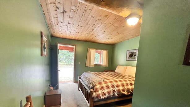 Une chambre avec un plafond en bois, un lit recouvert d’une couverture à carreaux, une fenêtre avec des rideaux et une porte donnant sur l’extérieur.