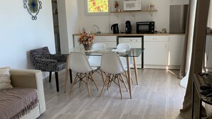 A modern living room with a sofa, a glass dining table with chairs, and a kitchen area with a microwave, coffee maker, and refrigerator. - Charmant T2 vue mer (Bastia)