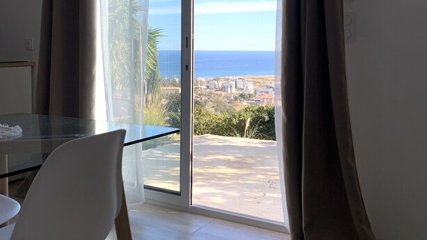 A room with a glass table and chairs, a sliding glass door leading to a balcony with a view of the sea and a cityscape. - Charmant T2 vue mer (Bastia)