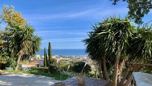 A wooden table with two glass candle holders, set on a patterned outdoor patio with a view of the sea and buildings in the distance. - Charmant T2 vue mer (Bastia)