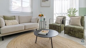 Una sala de estar moderna con sofá, sillón y mesa de centro. Hay una ventana con persianas, una obra de arte enmarcada en la pared y una planta en una mesita auxiliar.