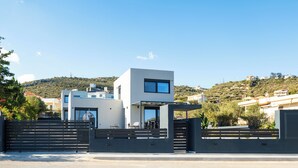 A modern two-story house with a gated entrance, a balcony, and a view of a hilly landscape.