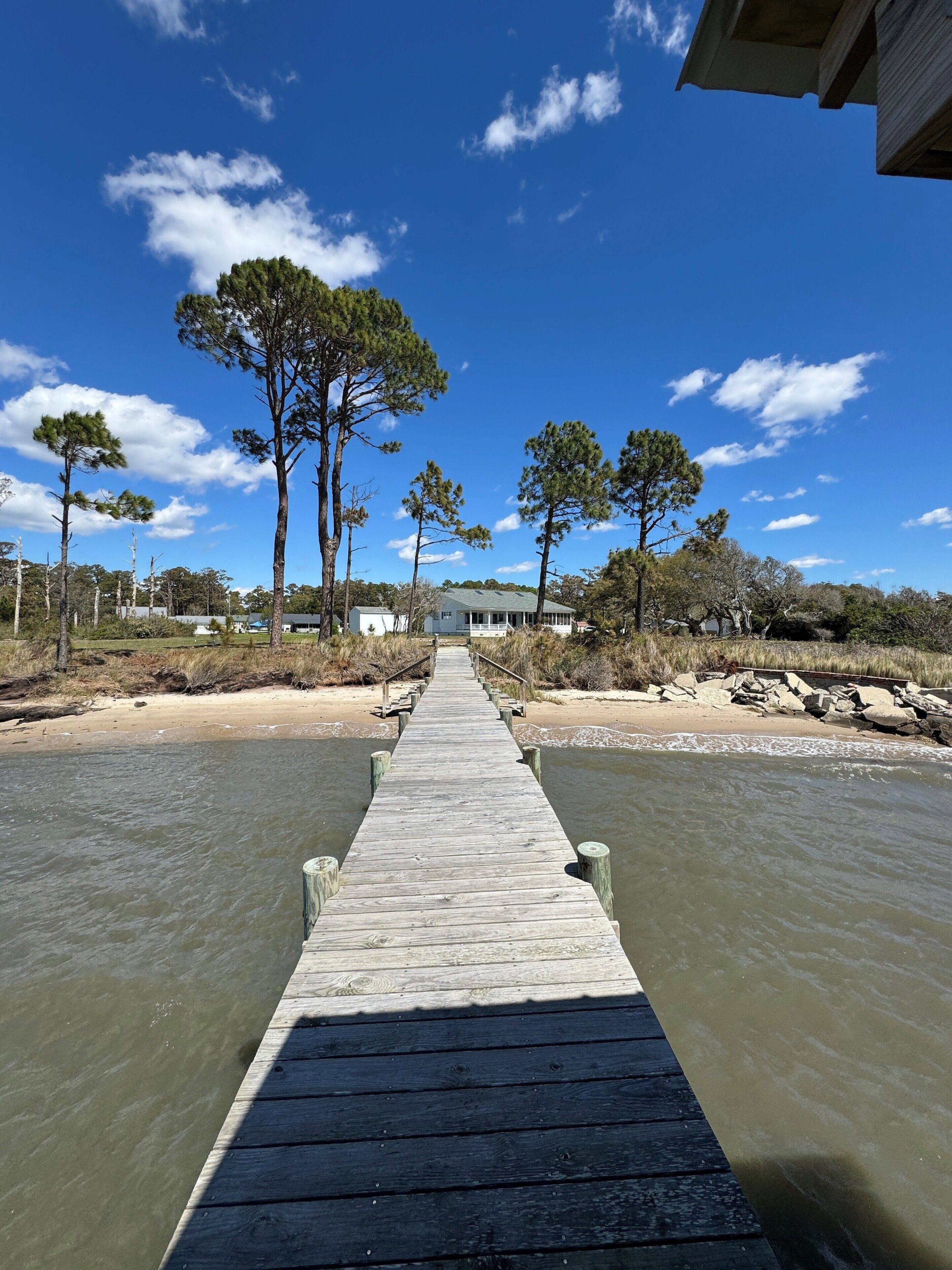 A wooden pier leading to a house with a porch, surrounded by trees and a clear sky.