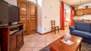A living room with a television, a sofa, a coffee table, and a chair. There is a kitchen area with wooden cabinets and a window with curtains. - Nice apartment for 4 people with WIFI, TV and pets allowed (Prè-saint-didier)