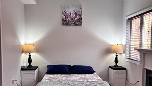 A bedroom with a bed, two bedside tables, a painting, a fireplace, and a window with blinds. - Beautiful Queen Room (Brampton)