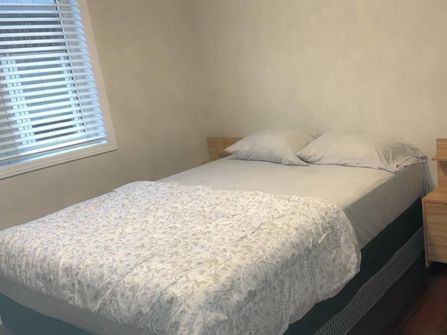 Een eenpersoonsbed met een bedsprei met patroon, een nachtkastje met lades, een zwarte koelkast en een raam met zonwering.