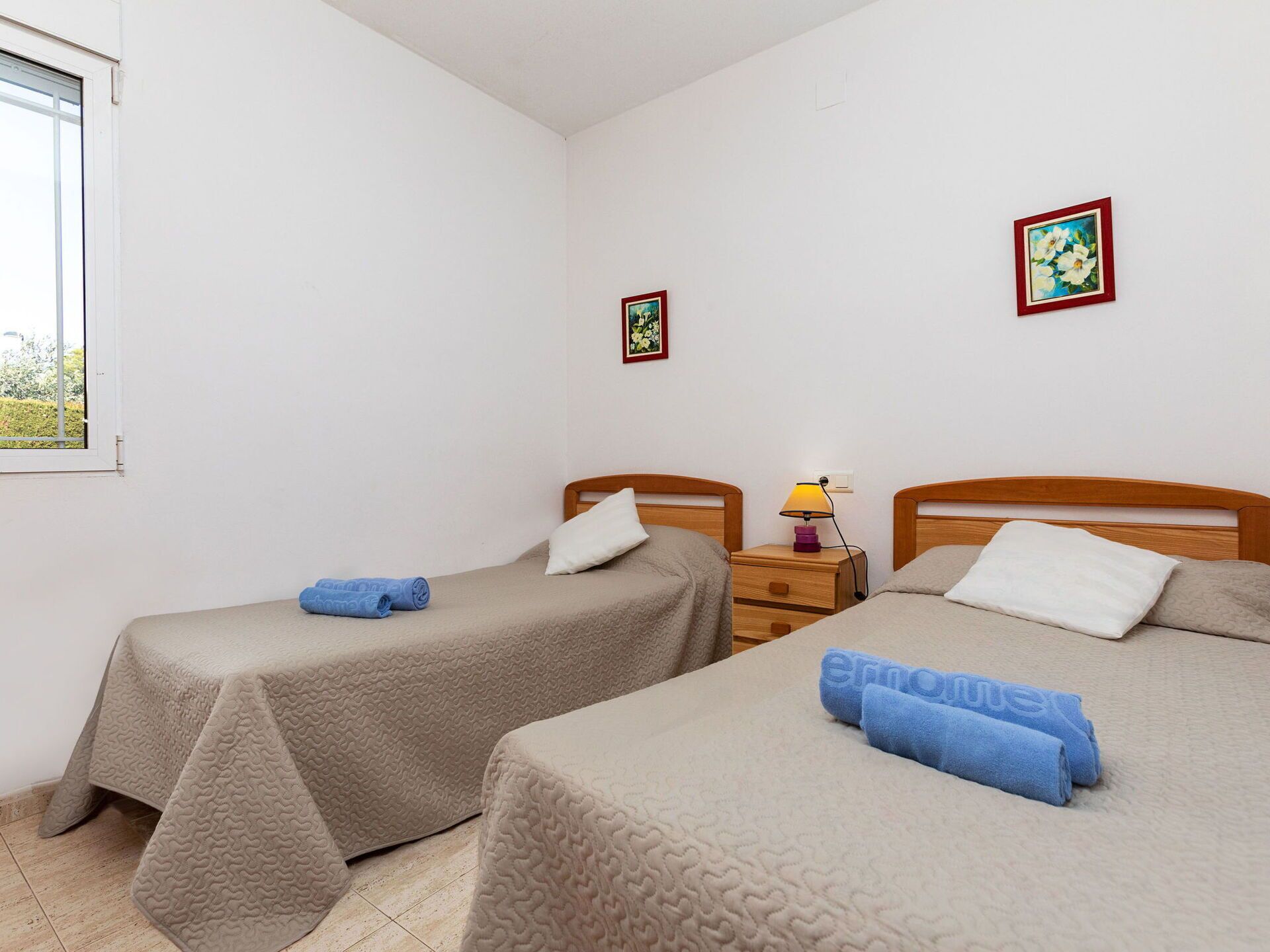 Une chambre avec deux lits simples, chacun équipé d’un oreiller et d’une serviette enroulée. Une table de chevet avec une lampe se trouve entre les deux lits.