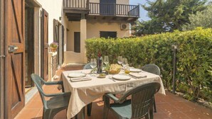 An outdoor dining area with a table set for two, surrounded by greenery and a building with a balcony.