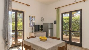A dining area with a table, chairs, a refrigerator, a TV, and a window with a view of greenery.