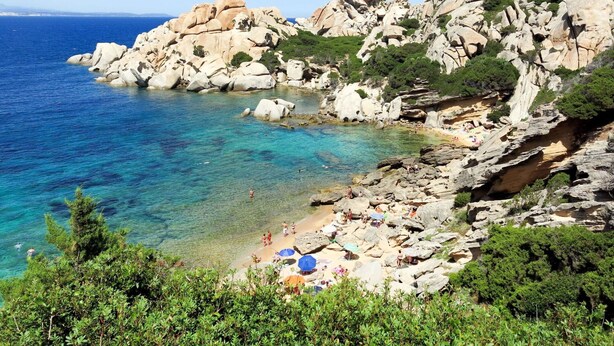 A rocky coastal area with clear blue water, rocky terrain, and greenery.