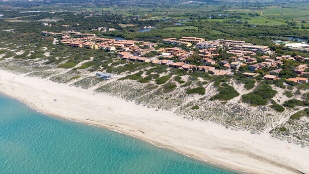 Vue aérienne d’une station balnéaire avec une plage, des bâtiments à étages et une végétation luxuriante environnante.