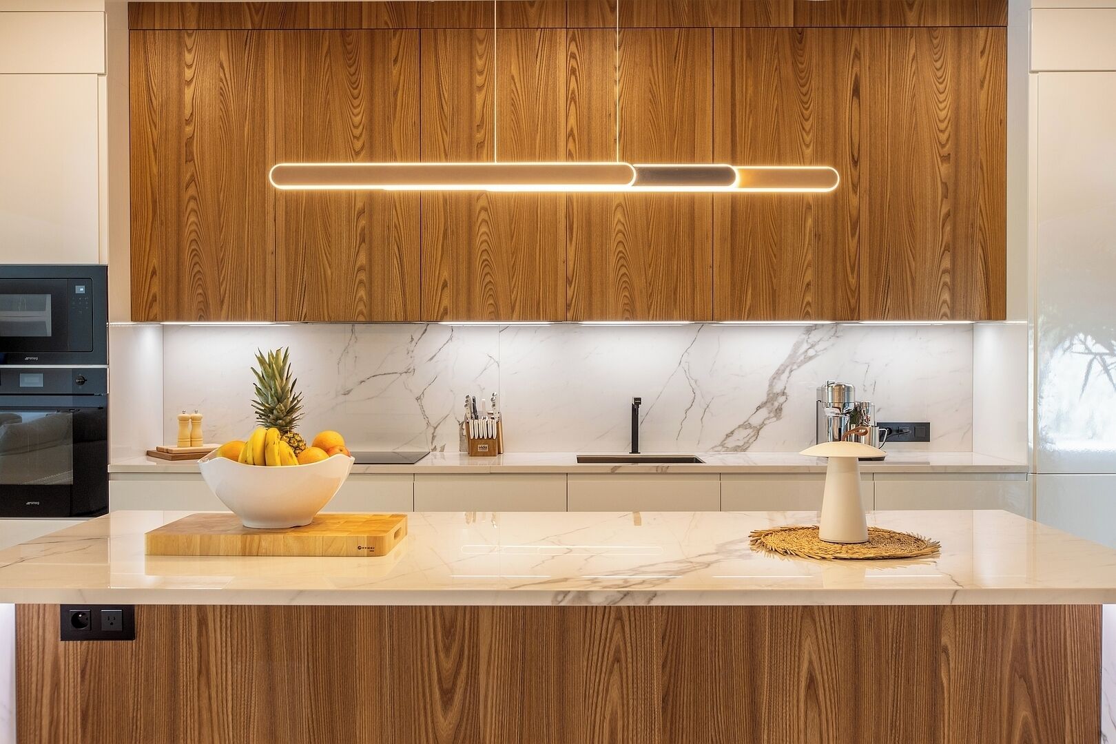 A modern kitchen with wooden cabinets, a marble backsplash, and a central island with a fruit bowl.