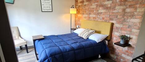 Un dormitorio con cama, mesita de noche, silla, estante en la pared y pared de ladrillo.