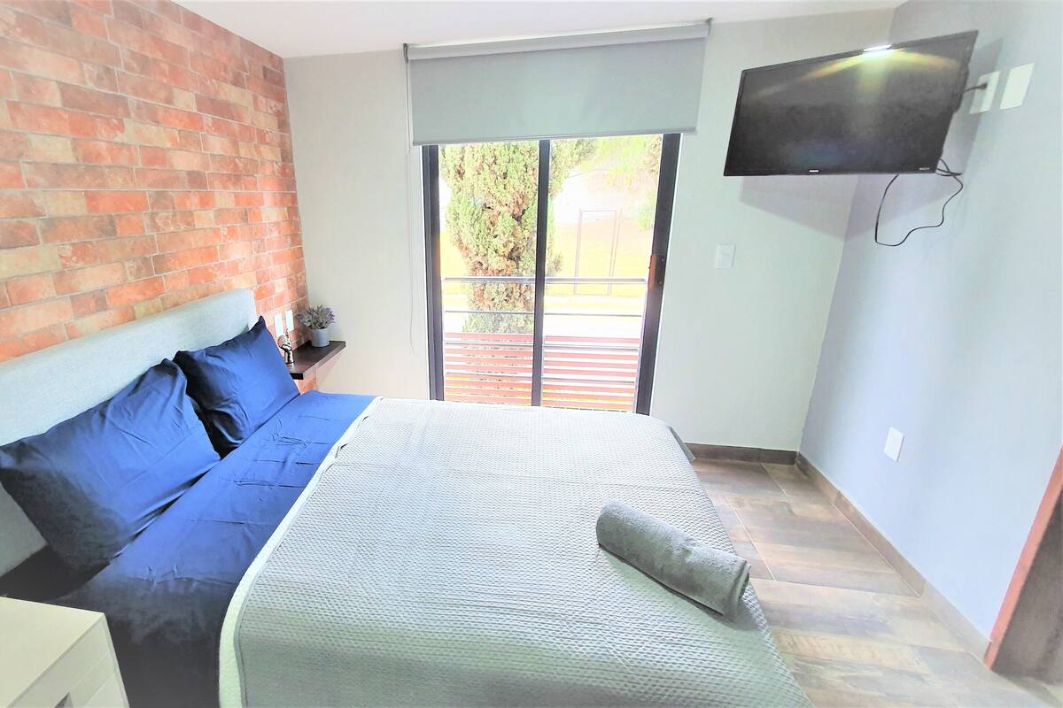 A bedroom with a brick wall, a bed with blue bedding, a TV on the wall, and a window with a view of greenery.
