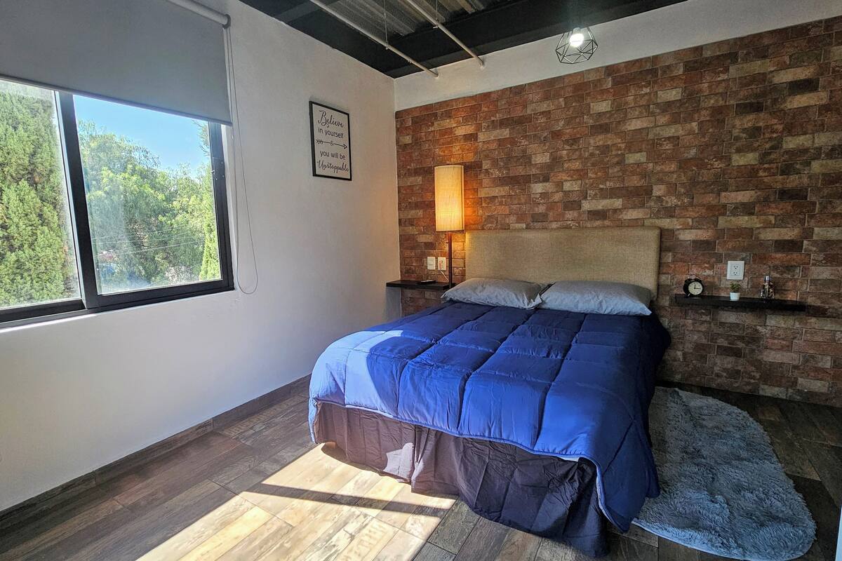 A bedroom with a large window, a brick wall, a bed with blue bedding, a bedside lamp, and a small shelf with a clock and a plant.
