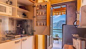 A compact kitchen with a built-in stove, a microwave, and a toaster. A small dining area with a table and chairs is visible through an open door, leading to a balcony with a scenic mountain view. - Beautiful apartment for 4 guests with hot tub, WIFI, TV, balcony and pets allowed (Vendrogno)