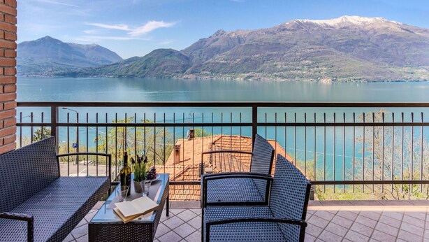 A balcony with wicker furniture, a table with a bottle and glasses, and a view of a lake with mountains in the background. - Beautiful apartment for 4 guests with hot tub, WIFI, TV, balcony and pets allowed (Vendrogno)
