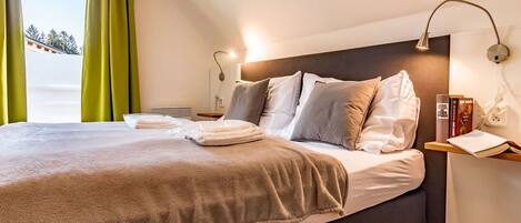 A neatly made bed with a brown blanket, a bedside table with a lamp, and a window with green curtains.