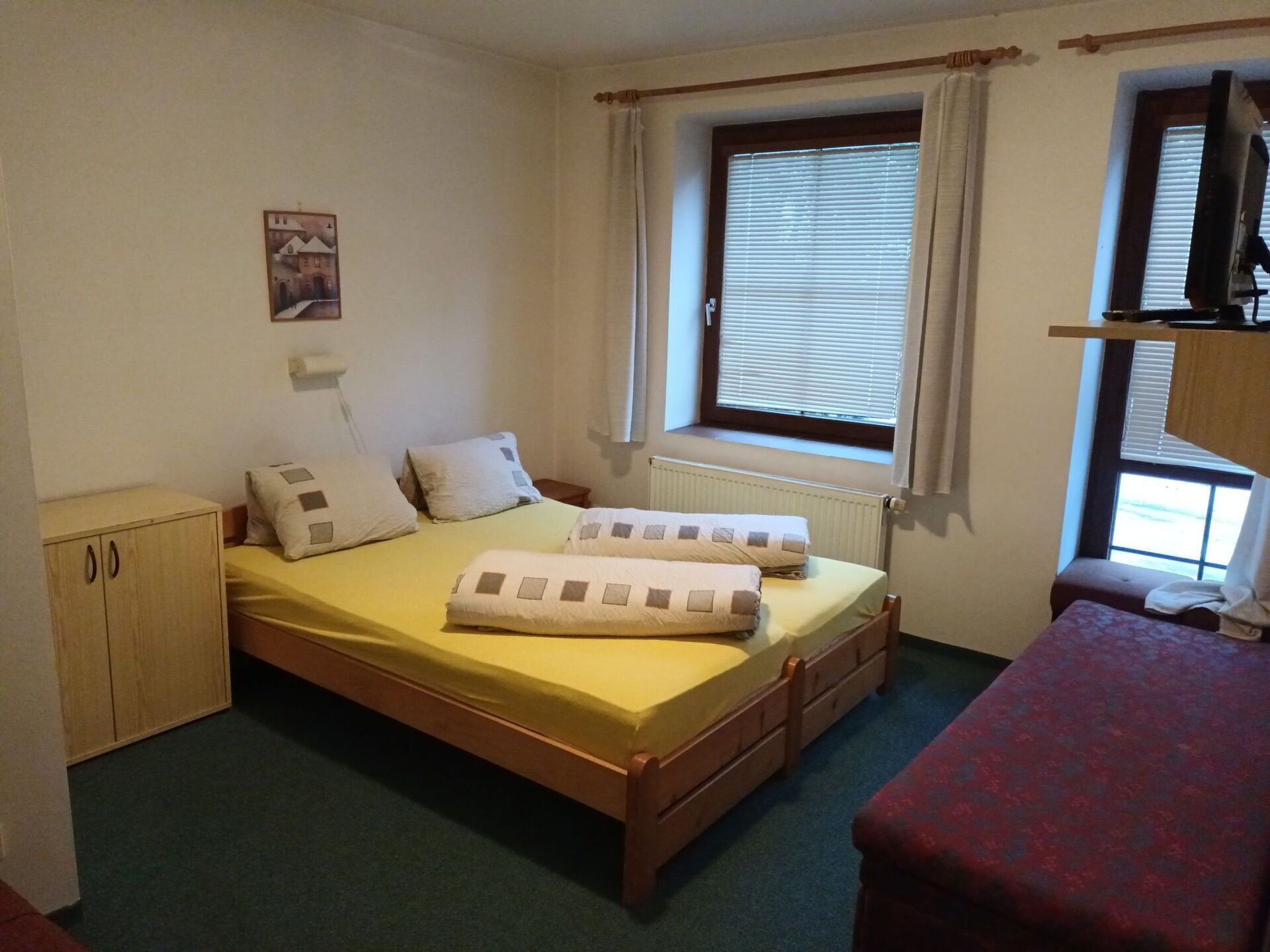 A small, tidy bedroom with a single bed, a wooden wardrobe, a window with blinds, and a television on a shelf.