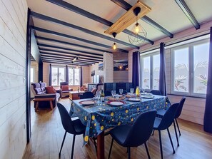 Un comedor con una mesa dispuesta para cuatro, vigas de madera en el techo y ventanas amplias decoradas con plantas.