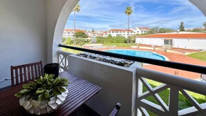 A balcony with a view of a swimming pool, a wooden table with a plant, and a white railing.