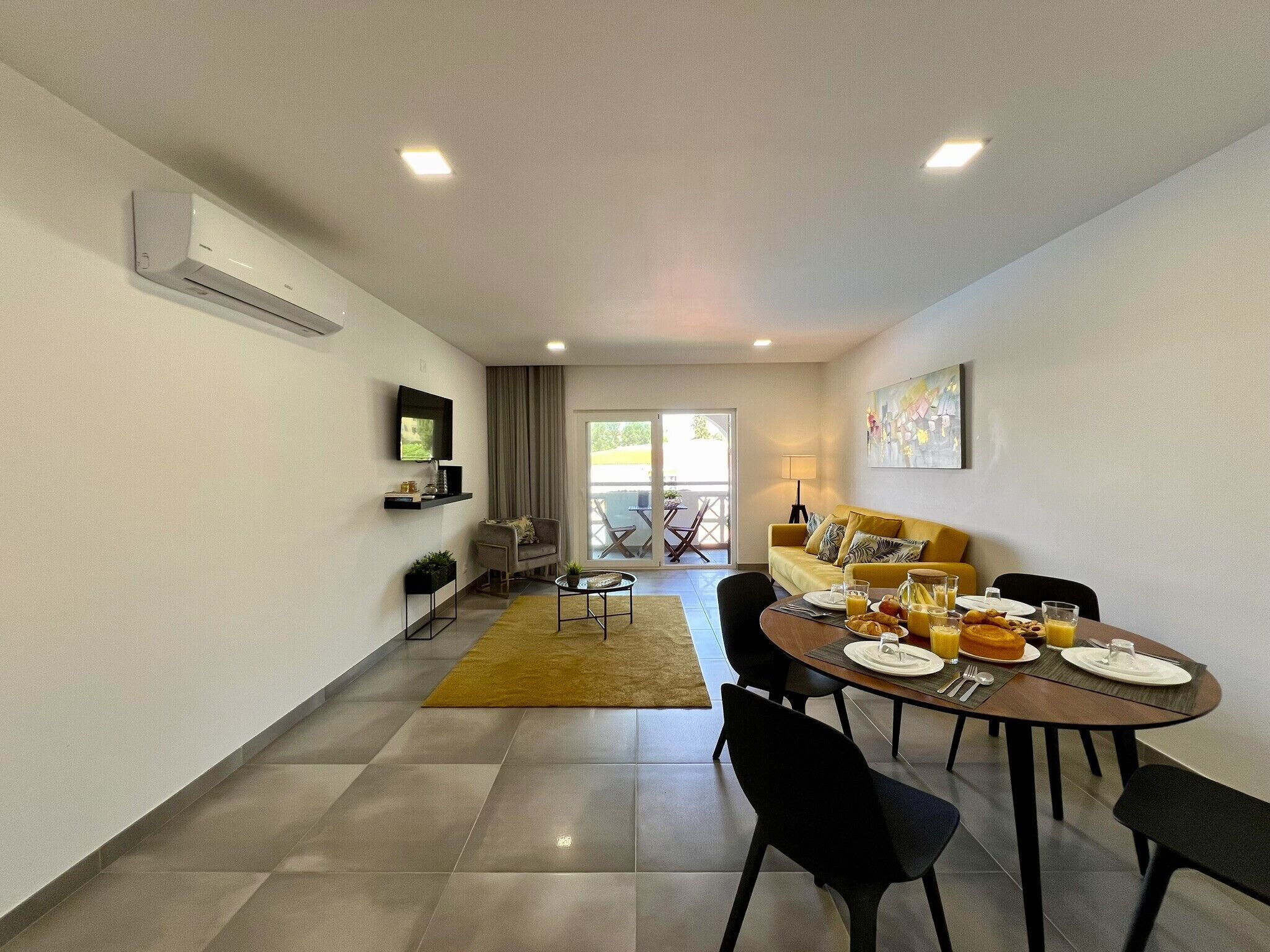 A modern dining area with a round table, chairs, and breakfast items. There is a sofa and a TV mounted on the wall.