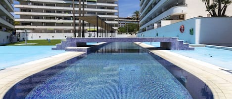 En swimmingpool med flisekant, omgivet af en lav mur og en bygning med flere balkoner i baggrunden.