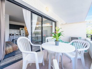 A balcony with a round table and white plastic chairs, a potted plant, and a sliding glass door leading to an indoor area with a bed and a chair.