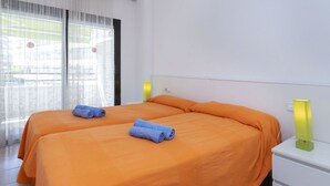 Camera d'albergo con due letti, biancheria arancione, un comodino bianco e una finestra con tende leggere.