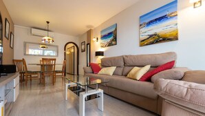 Una sala de estar moderna con una mesa de centro de vidrio, un sofá color beige con cojines rojos y amarillos, y dos cuadros en la pared.