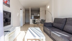 Een moderne woonkamer met een grijze bank, een witte salontafel en een televisie op een witte kast. Er is een eethoek met een tafel en stoelen, en een keuken op de achtergrond.